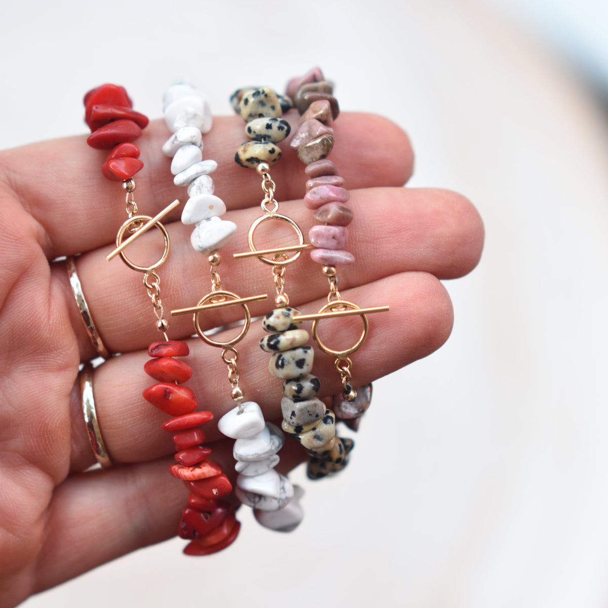 Four beaded toggle bracelets held in a hand. The bracelets feature chunky red coral-like beads, smooth white beads, patterned gray and black beads, and dusty pink beads, all with gold-toned toggle closures.