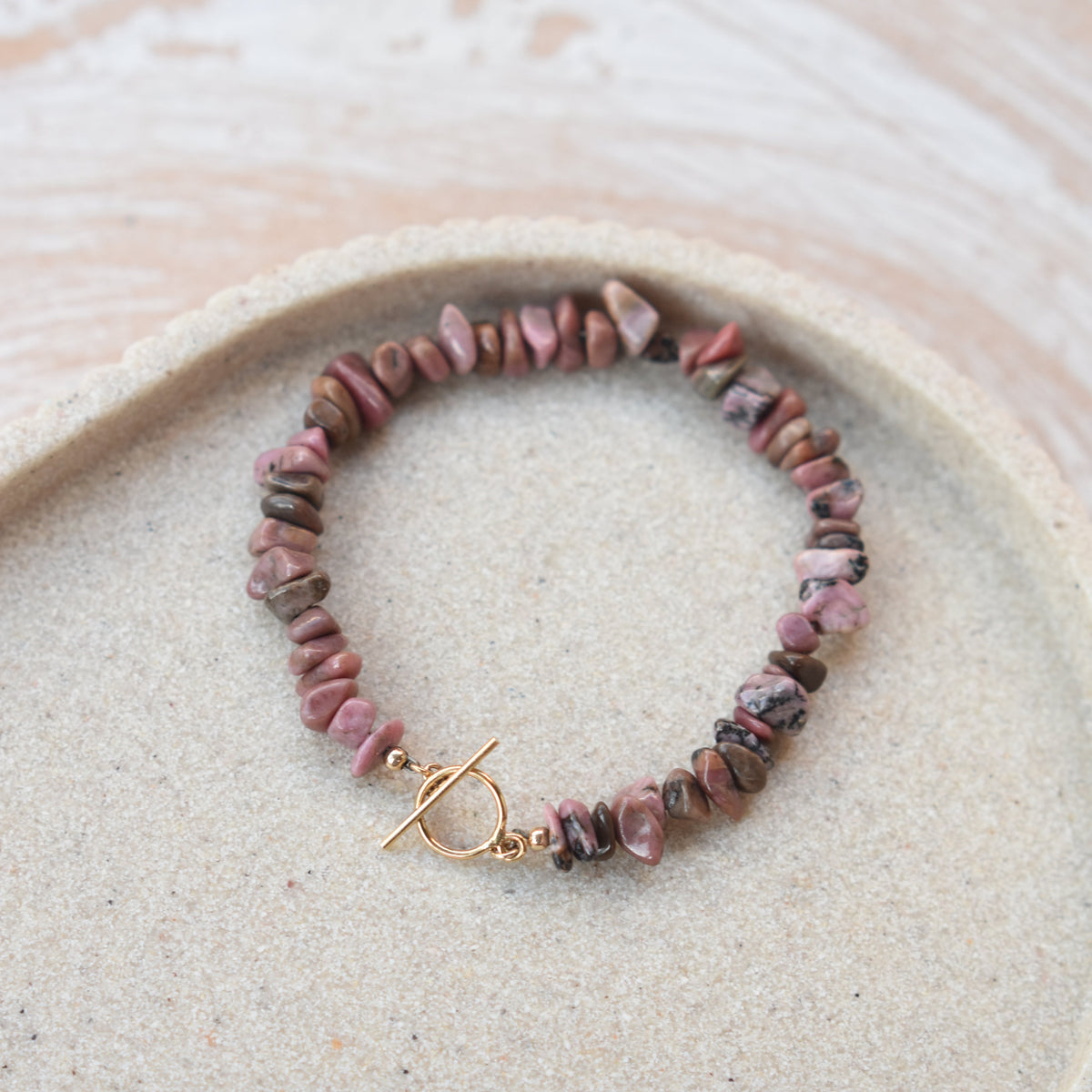 A beaded bracelet made of small pink and brown stone chips with a gold toggle clasp, displayed on a beige textured tray.