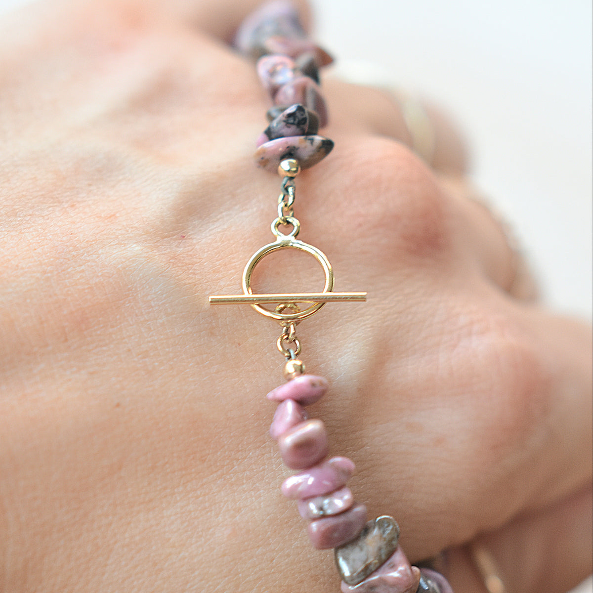 A close-up of a hand wearing a beaded bracelet made of pink, purple, and gray stone chips with gold-toned metal beads and a toggle clasp.