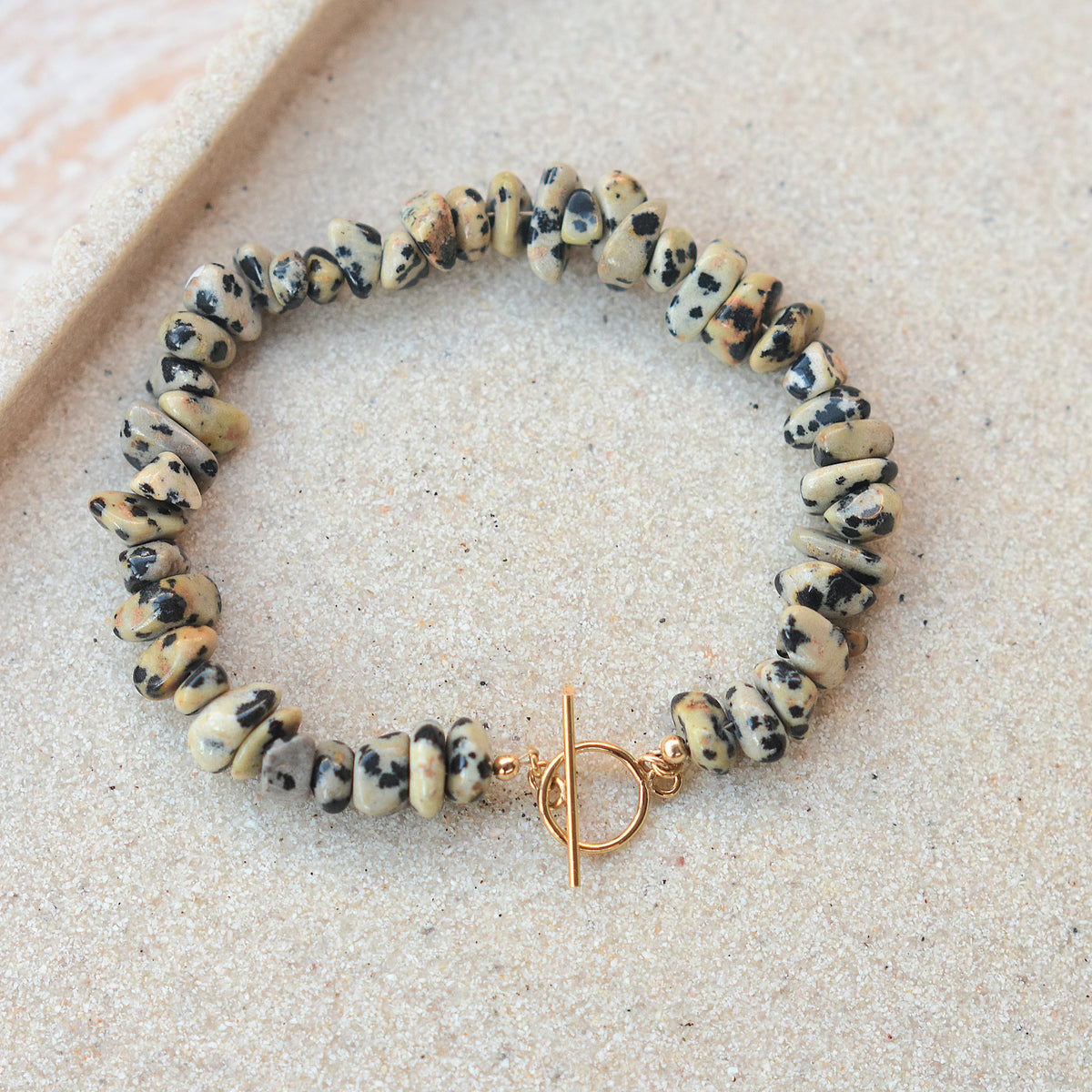 A beaded stretch bracelet made of irregular cream and black speckled stones, featuring a gold-tone toggle clasp, displayed on a light beige textured surface.