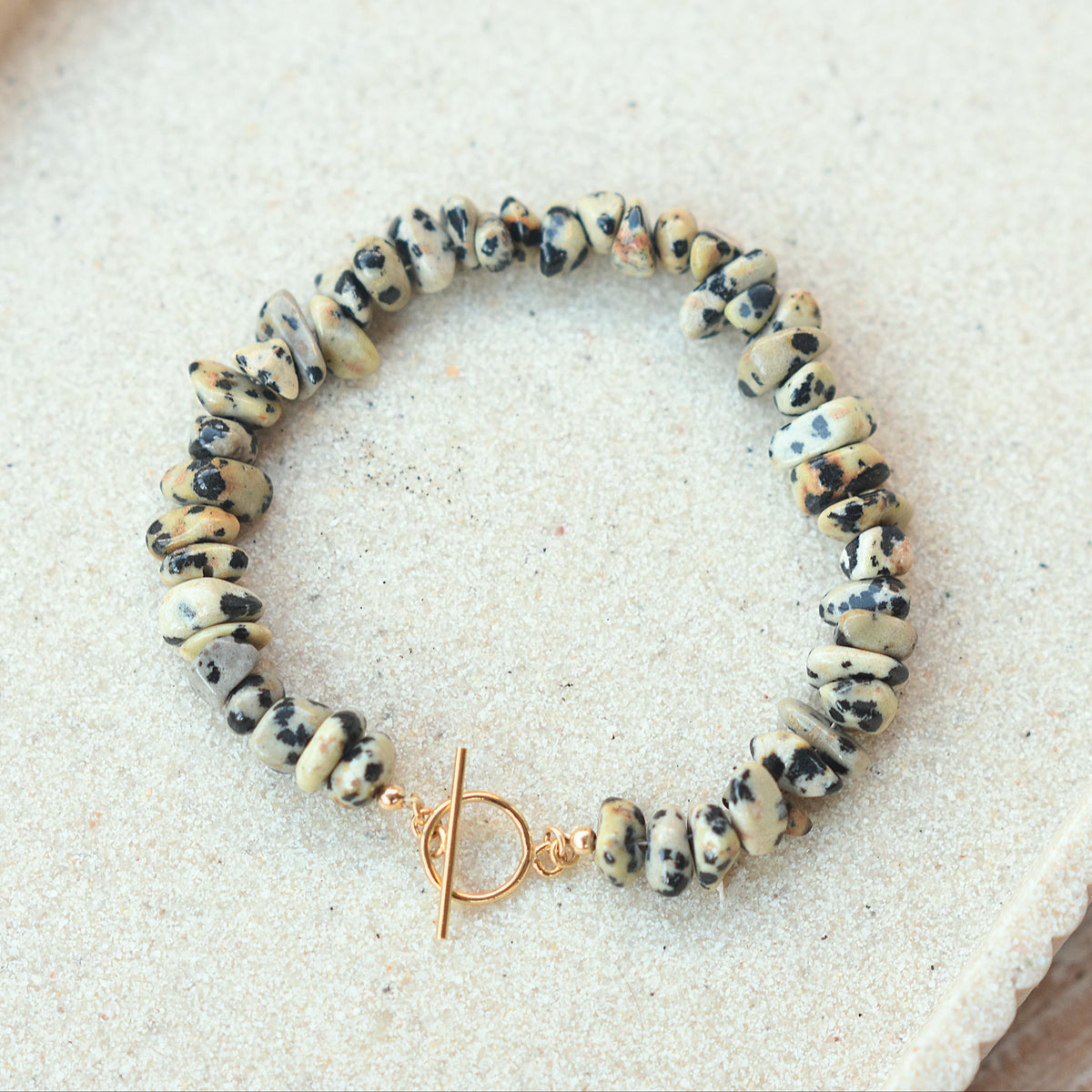 A beaded stretch bracelet made of irregular black and white marbled beads with a gold toggle clasp, displayed on a textured beige surface.