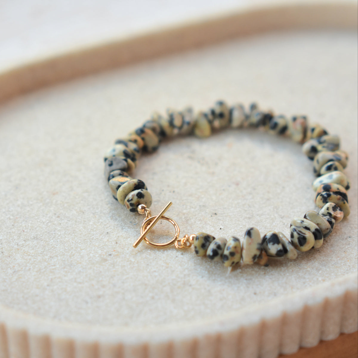 A beaded stretch bracelet with marbled black, white, and beige beads and a gold toggle clasp, displayed on a textured beige surface.