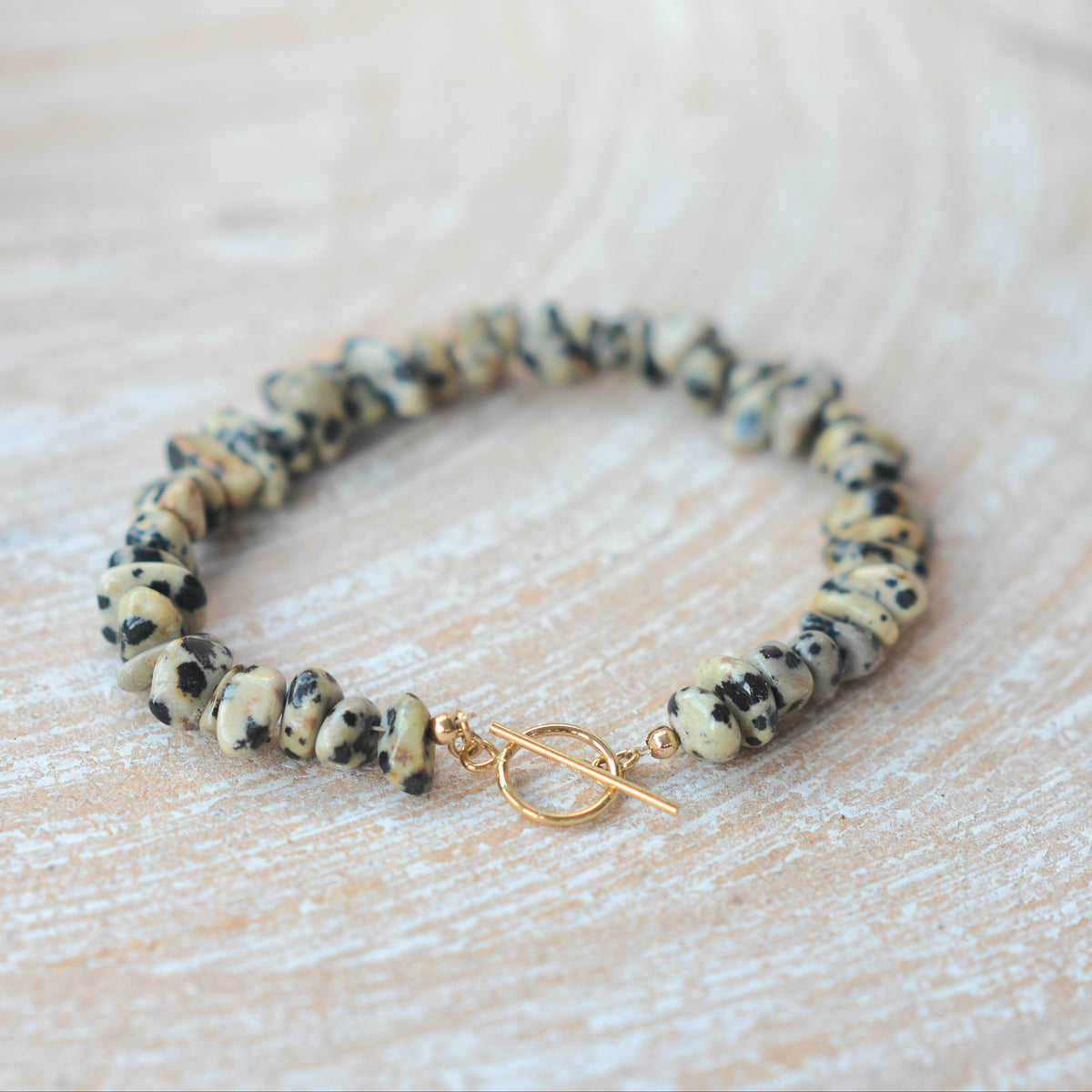 A beaded stretch bracelet made of irregular natural stone beads in beige, black, and cream, featuring a gold toggle clasp, resting on a light wood surface.