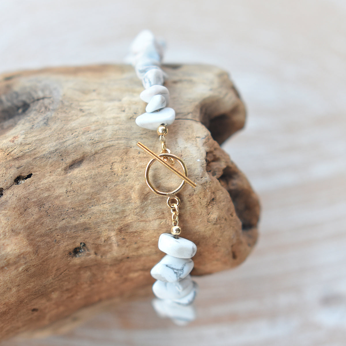 A delicate bracelet made of white stone chips and gold-toned metal, displayed on a piece of driftwood against a light, softly blurred background.