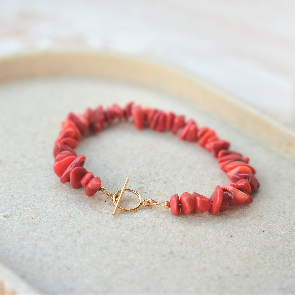 A red chunky stone bead bracelet with a gold toggle clasp, displayed on a textured beige tray.