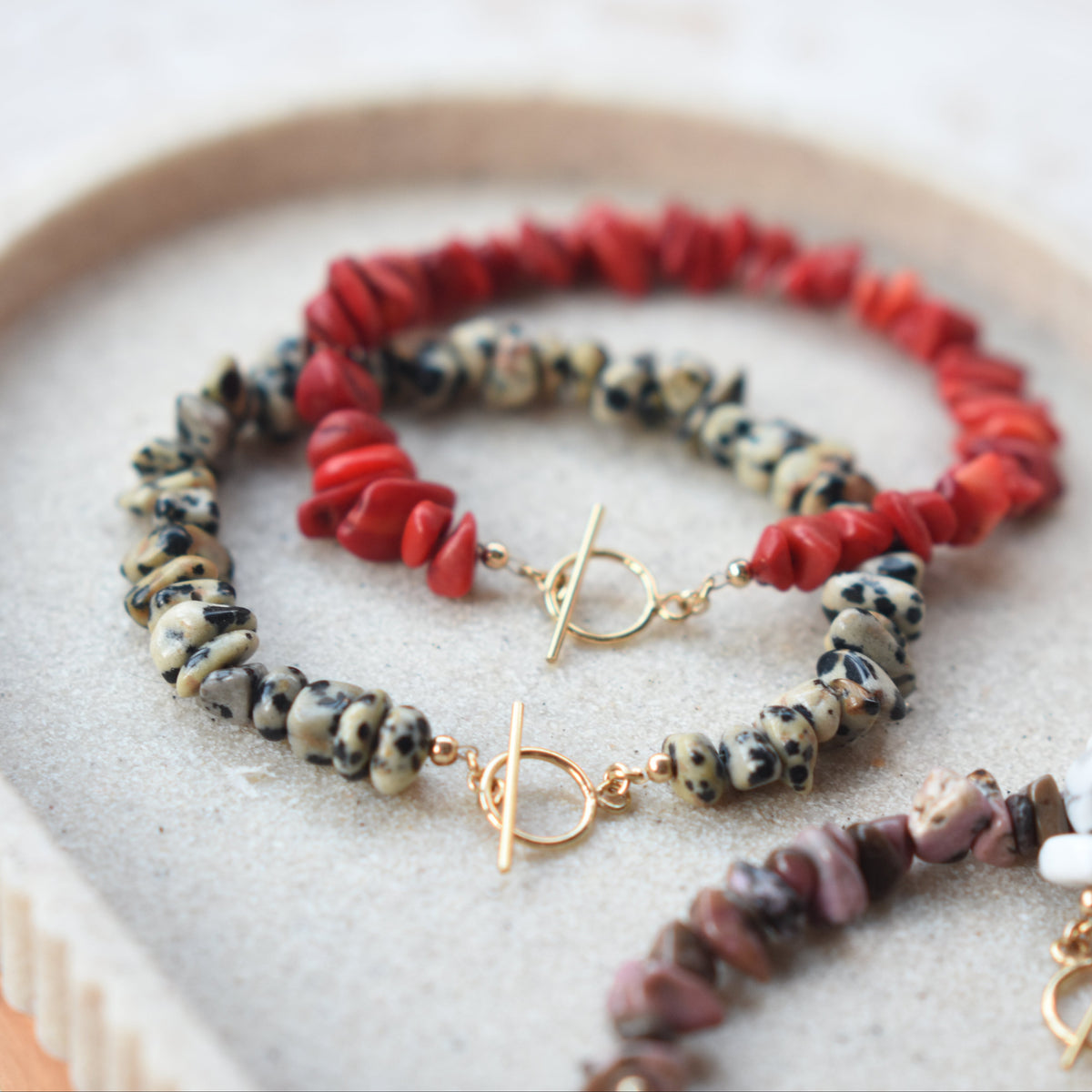 Three beaded stretch bracelets with toggle clasp rings, displayed on a beige tray. The bracelets feature red chips, black-and-white mottled chips, and pink-and-white mottled chips, all with gold-toned hardware.