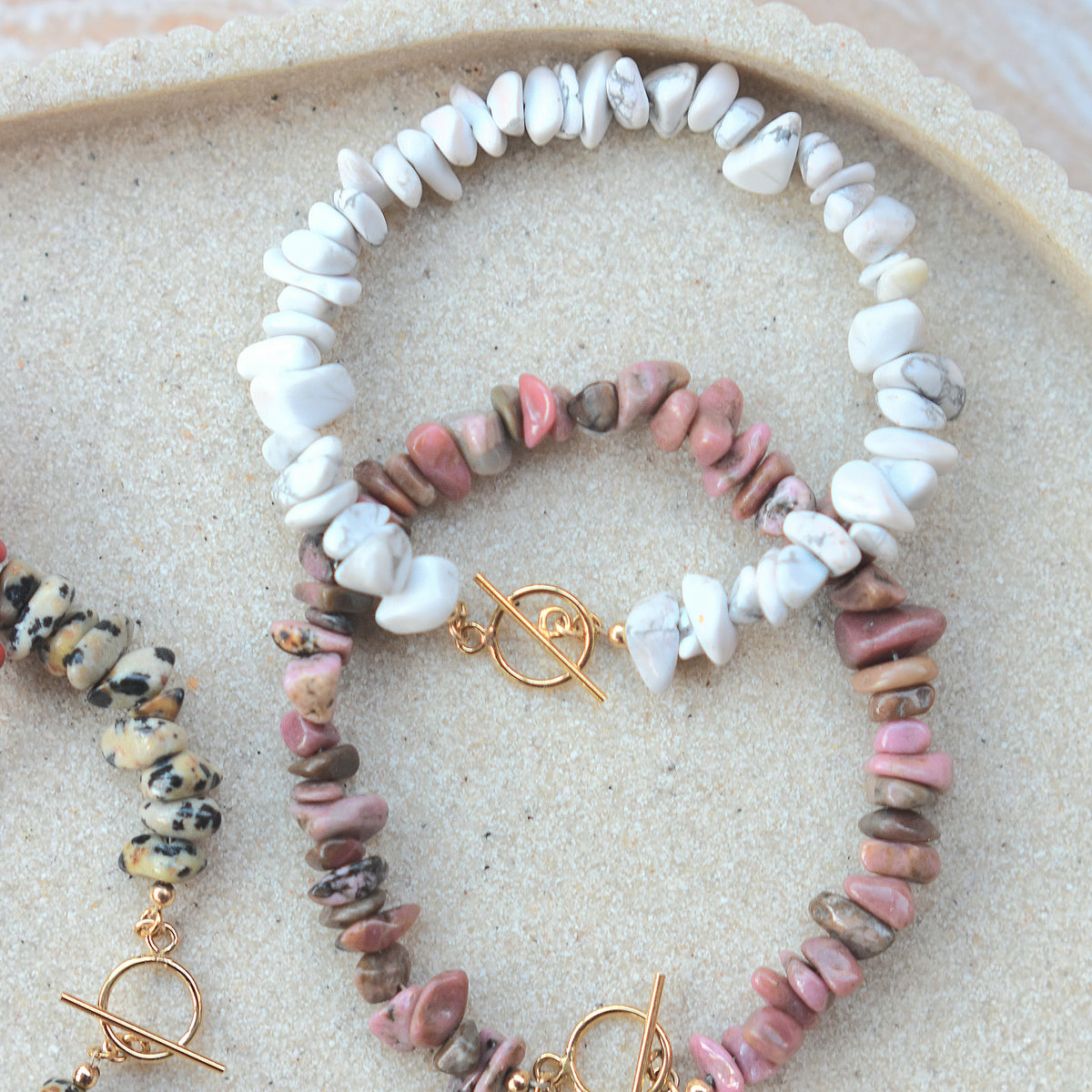 A set of handmade beaded bracelets displayed on a scalloped beige tray. The bracelets feature natural stone chips in white, pink, brown, black, and coral red, with gold-toned toggle clasp closures.