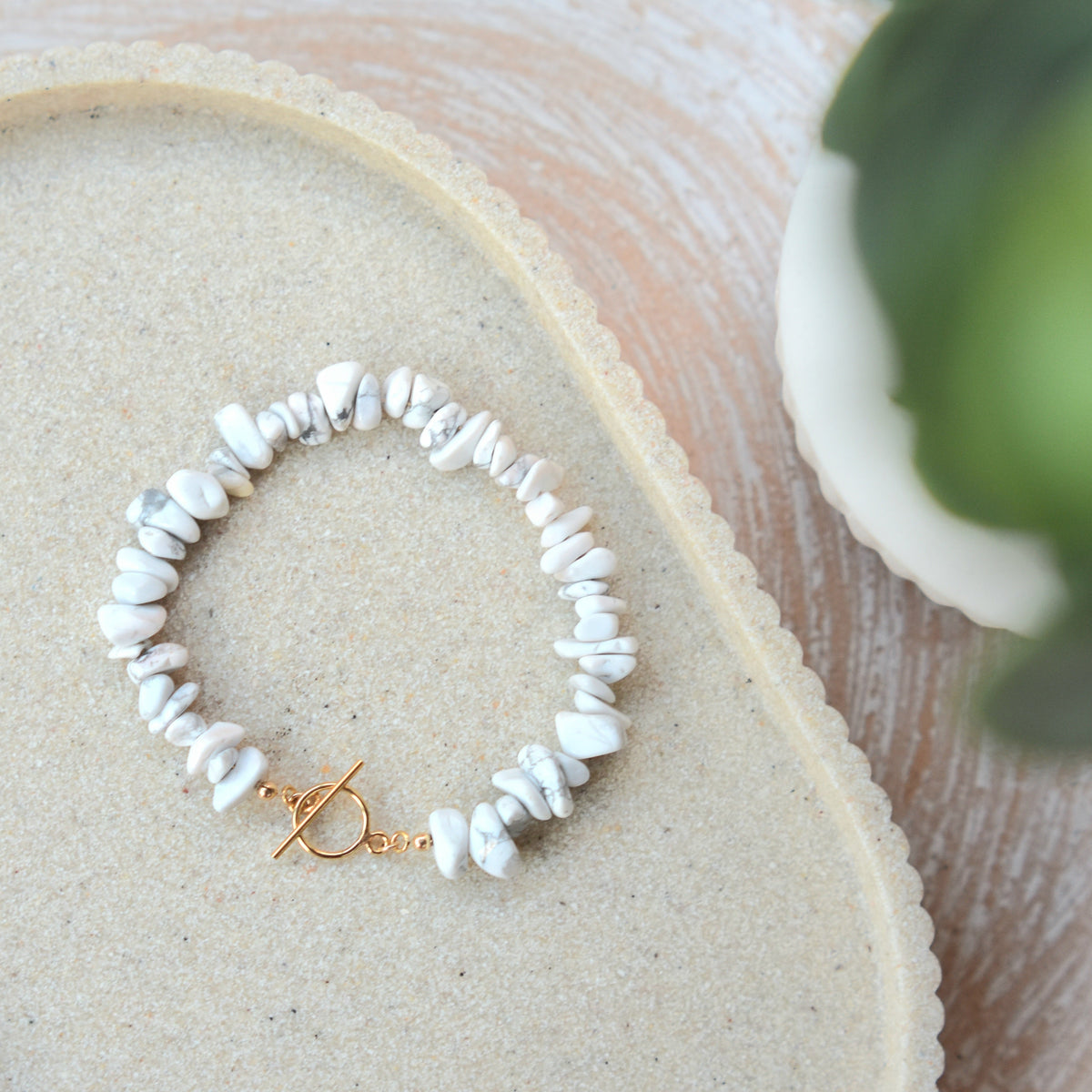A delicate bracelet made of small white shell chips with a gold toggle clasp, displayed on a beige textured tray.