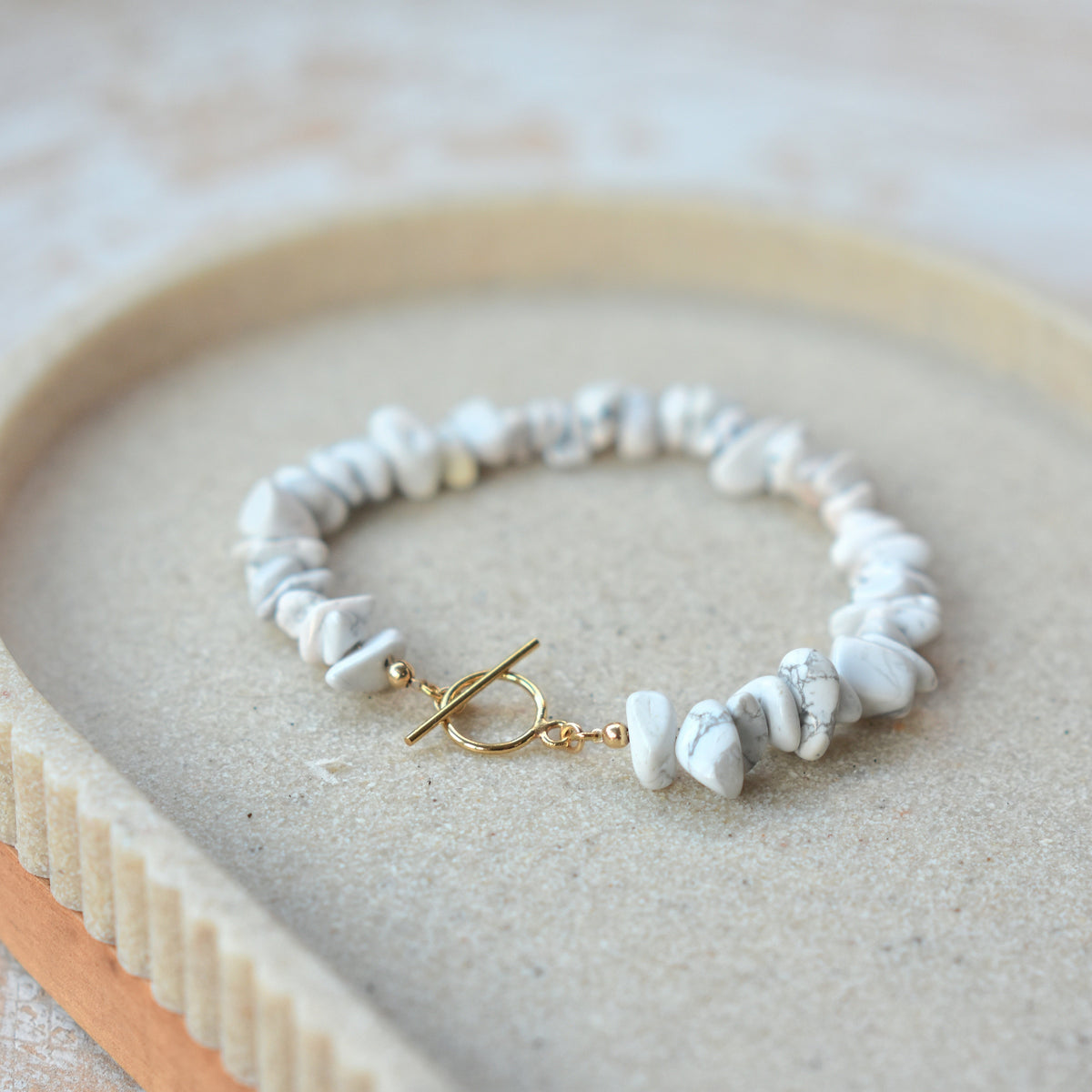 A beaded bracelet made of irregular white stone chips with a gold toggle clasp, displayed on a textured beige surface inside a light wood tray.