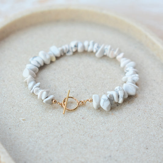 A delicate bracelet made of small white and gray stone chips with a gold toggle clasp, displayed on a beige textured surface inside a light wood tray.